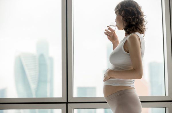 Side view of young pregnant model drinking water and looking at city scenery in the window. Future mom expecting baby relaxing at home with glass of drink. Copyspace